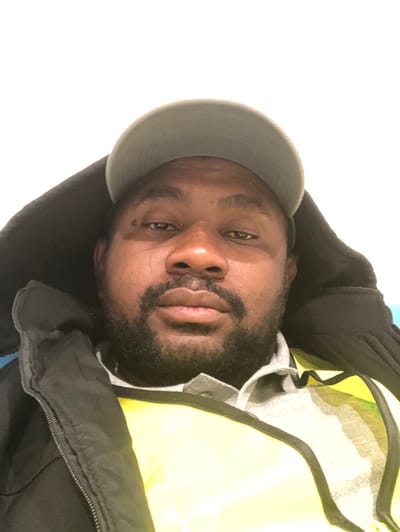 A man with a beard and wearing a gray baseball cap looks directly at the camera. He is also wearing a black jacket and a yellow high-visibility vest. His head is slightly tilted to the side. The background is primarily bright white on the left. The lighting is flat.