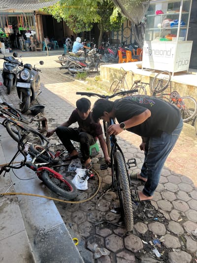 In Keude Ulim, Indonesia, on a sunny day, two young men are working on bicycles outside a shop. The scene is set on a paved area in front of a building with a covered awning and a display case. In the foreground, various bicycle parts and tools are scattered on the ground, along with several bicycles in various states of repair. One man, wearing a dark t-shirt and dark pants, is bent over a bicycle, appearing to be making adjustments to the wheel. He is sitting on a small green stool. The second man, dressed in a black t-shirt and blue jeans, is also working on a bicycle, leaning over it and looking at the frame. Several motorcycles are parked nearby. The background shows a street with more shops and people.