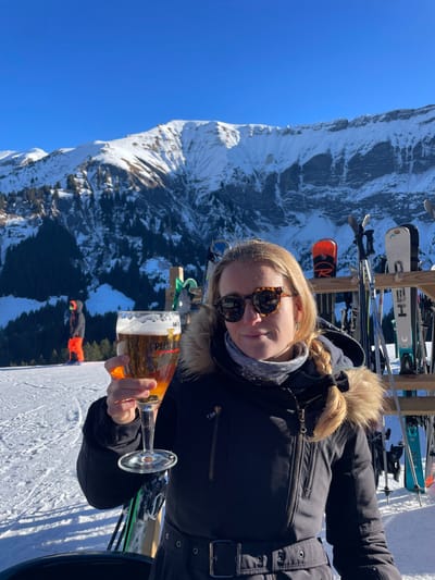 An outdoor scene captured on a clear, sunny winter day in Megève, France, features a blonde female in the foreground, positioned slightly to the right. She wears a dark winter jacket with a fur-lined hood, a grey patterned neck gaiter, and tortoiseshell sunglasses, displaying a slight smile. Her right hand holds a stemmed glass containing a golden-amber beer with a foam head, bearing the "Pelforth" brand label. To her immediate right, a wooden rack stores multiple pairs of skis and poles, some identifiable by brands such as Rossignol and Head. In the mid-ground to the left, another individual, clad in a dark jacket and bright orange ski pants, stands on a snow-covered slope, facing away from the camera. The background is dominated by a vast, snow-capped mountain range featuring rugged peaks, exposed rock faces, and coniferous tree lines under a bright, cloudless blue sky.