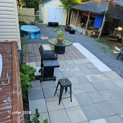 The image depicts an outdoor backyard scene in Saint-Calixte, Canada, on July 11, 2026. The primary focus is a paved patio area constructed with square interlocking stones in varying shades of grey and beige. A black gas grill stands on the patio, to the right of a metal stool with a circular seat. Adjacent to the patio is a wooden deck with weathered red paint. A collection of metal grilling tools, including two long-handled forks, are laid on the deck.
In the background, a small, round, inflatable blue and orange kiddie pool is partially filled with water. Behind the pool, a light yellow shed with double white doors is visible. Further back, a rustic, open-sided covered structure with a corrugated metal roof is present, supported by wooden posts. A blue towel is hanging from a rail within this structure, and lawn chairs are also visible. The ground surrounding the patio and shed is covered with gravel. Green foliage, including a tall weed growing near the patio and a bush to the left, is present throughout the scene. The overall impression is one of a functional but somewhat unkempt outdoor living space.