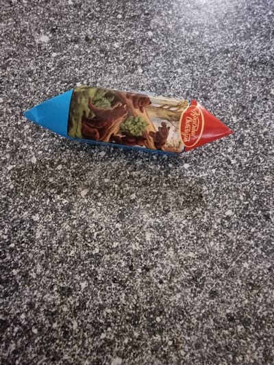 A colorful candy wrapper featuring a forest scene with bears is placed on a speckled grey surface, possibly a countertop or floor. The wrapper is pointed at both ends, with a blue section on the left and a red section on the right. The central part of the wrapper depicts a mother bear and her cubs in a wooded area. On the red section, there is text in Cyrillic. The image does not provide any information about the location in Belozyorny, Russia.