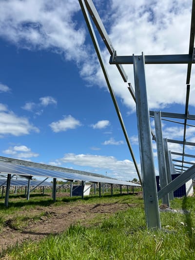 The image shows a field in Grange Ville, Ireland, with rows of solar panels installed. The panels are mounted on a metal framework. The foreground shows the supporting metal framework with a view of the sky with scattered clouds. The ground is covered in grass and dirt, indicating recent construction or maintenance. No people are visible in the frame. The scene presents a sunny day with a clear blue sky interspersed with fluffy white clouds. The image is taken from a low angle, looking upwards.