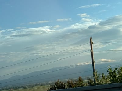 The image shows a landscape under a partly cloudy sky, likely taken during the daytime. The scene is dominated by a vast expanse of sky filled with streaky white and gray clouds against a pale blue backdrop. In the foreground, a wooden utility pole with insulators stands prominently, with several dark power lines stretching horizontally across the frame.
Below the sky and power lines, a hazy mountain range is visible in the distance. The mountains appear layered, with some peaks showing soft, misty outlines. In the valley between the viewer and the mountains, there's evidence of a settled area with greenery and what looks like agricultural fields. The overall impression is one of a rural or semi-rural setting.
The presence of utility poles and power lines suggests human infrastructure within this natural landscape. The lighting indicates it is likely daytime, possibly late afternoon given the soft light and shadows, although it's difficult to be precise without more cues. The weather appears mild, with clouds suggesting a dynamic sky but not necessarily inclement conditions. There are no visible people, text, or specific identifiable landmarks that would pinpoint an exact location beyond the general mountainous terrain. The scene evokes a sense of open space and a connection to nature, interspersed with signs of human development. The crops provided offer closer views of the sky, clouds, and the detail of the utility pole and wires.