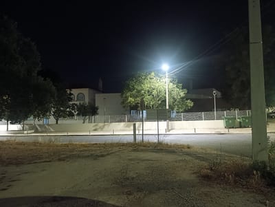 The image captures an outdoor scene at night in Glória do Ribatejo, Portugal. In the foreground, a dirt and gravel area is visible, with dry brush and sparse vegetation. A paved road runs horizontally across the mid-ground, bordered by a sidewalk. Behind the sidewalk, a white, modern-looking building with arched windows and a red-tiled roof is situated. Several trees with dense foliage are positioned in front of and around the building.
Illumination is provided by a bright streetlamp in the center of the frame, casting a strong light on the surrounding trees and area. In the background, a tall, light-colored concrete utility pole stands on the right side of the image. Two large, green dumpsters are visible on the sidewalk near the building. The sky is dark, indicating nighttime. The overall atmosphere is calm and still, with no people or discernible activity.