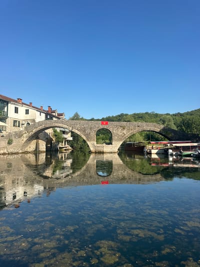 The image captures a picturesque view of the Prince's Bridge (Most Kneza Danila) in Rijeka Crnojevića, Montenegro. The stone bridge, with its characteristic arches, spans a calm river, reflecting the clear blue sky and the surrounding architecture. On the left side of the bridge, buildings with white facades and red-tiled roofs are visible, hinting at the presence of a town. A red flag, possibly the Montenegrin flag, is prominently displayed on the central arch of the bridge.
Several boats are moored along the right bank of the river, suggesting a popular tourist spot for river excursions. The water in the foreground is shallow and clear, allowing the viewer to see the riverbed covered with aquatic vegetation and submerged rocks. The overall atmosphere is serene and tranquil, bathed in bright daylight. The dense green foliage on the distant hills provides a lush backdrop to the scene.
