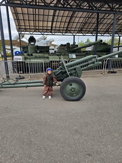 The image shows a boy in a blue hat, brown jacket, and pants standing next to a green artillery piece at the Vadim Zadorozhny Technology Museum in Arkhangel'skoye, Russia. The artillery piece is positioned on a paved surface near a covered structure with a black metal frame. An aircraft is partially visible under the structure. A metal fence separates the artillery piece from the paved area.