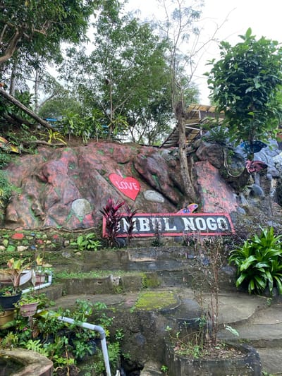 The image shows a large, painted rock formation in Kecamatan Manyaran, Indonesia, at the coordinates -7.826 latitude and 110.816 longitude. The rock is painted in various shades of red, pink, and green. A red heart with the word "LOVE" written inside it is painted on the right side of the rock. A red and white sign is mounted beneath the rock, reading "MBUL NOGO" in white capital letters on a red background. The rock and sign are framed by lush greenery and a series of stone steps leading down to the bottom of the image. The scene appears to be a small outdoor space, possibly a garden or a park.