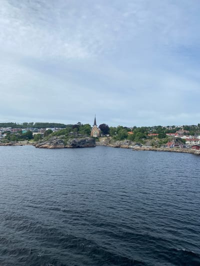 This is a landscape photograph taken from the water, looking towards a rocky coastline. The image is oriented vertically, with the sky at the left and the sea at the right. The coastline is dotted with buildings, many of which appear to be houses with red roofs, nestled among lush green trees. A prominent stone church with a spire stands on a cliff overlooking the water. The sea is a deep blue with visible ripples, suggesting a gentle breeze. The sky is overcast with soft, diffused light, indicating it is likely daytime, possibly late afternoon or early morning. The scene evokes a sense of tranquility and the picturesque beauty of a coastal town. The location is identified as Larvik, Norway, which aligns with the typical architecture and coastal landscape of Scandinavian regions. There is no visible text in the image.