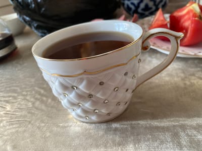 This close-up shot features a white teacup filled with dark tea, its surface adorned with a quilted pattern and sparkling rhinestones. A delicate gold trim encircles the rim and the handle of the cup. The teacup rests on a textured, light-colored tablecloth. In the background, slightly out of focus, are other elements of a table setting. To the left, part of a dark object, possibly a bowl or vase, is visible, along with a white dish. To the right, a pink-rimmed plate holds several pieces of sliced red fruit, likely tomatoes, hinting at a savory accompaniment to the tea. The lighting suggests it might be daytime, with soft illumination casting gentle highlights on the tablecloth and the cup's embellishments. The scene evokes a sense of quiet refreshment or a moment of leisure. No visible text is present in the image.