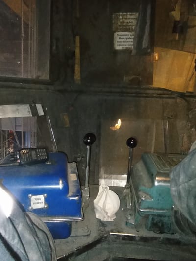 The image appears to be taken from inside a vehicle, possibly a train or industrial machinery, looking towards a control panel. The scene is dimly lit, suggesting it might be nighttime or an indoor setting.

In the foreground and center, two control levers with black spherical knobs are visible, suggesting active operation or preparation for operation. To the right of these levers, a blue control unit is prominent, with a small, white label attached by screws. This label has text in Cyrillic, which reads "Ход телега". Adjacent to this blue unit is a black handheld radio or communication device. Further back and slightly to the left, a teal-colored control unit is visible, also with a lever. A crumpled white piece of cloth or paper is resting on the surface near the teal unit.

On the left side of the image, a window reflects a dimly lit environment. Through the window, some text is visible on what appears to be a notice or instruction board. The text is in Cyrillic and partially legible, including phrases like "Машинист кра..." and "Перед работ...". These fragments suggest instructions or warnings related to the operation of the machinery. The overall impression is one of an operational or maintenance setting, with the focus on the control mechanisms of the vehicle. The limited visibility and focus on controls imply the image was taken during a specific operational phase or for documentation purposes. The reflection in the window suggests an indoor location, possibly a depot or industrial building. There are no visible people in the frame.