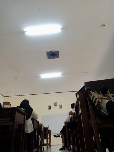 This image displays an interior classroom scene in Kawungluwuk, Indonesia. The white ceiling features two illuminated fluorescent tube lights and a central dark, square ventilation grille. Walls are painted light cream, and the floor is tiled. In the foreground right, a person's legs, clad in black pants and black-and-white striped sneakers, are propped on a wooden desk. To the mid-left, a student wearing a white shirt and black hijab is seated at a wooden desk, facing away. Another student, visible mid-right, wears a white shirt, dark pants, and black shoes with white socks, seated at a desk. A whiteboard and three framed portraits hang on the rear wall. Wooden desks and chairs populate the room, consistent with a scholastic environment.