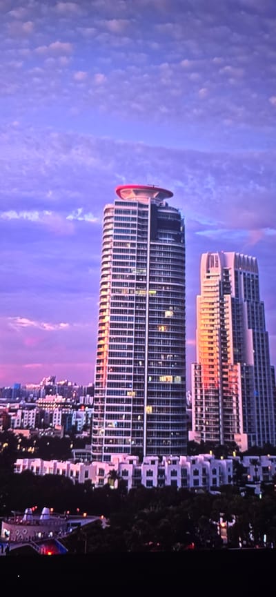 The image shows a cityscape at sunset with a purple and pink sky. Two tall, modern skyscrapers dominate the center of the frame, with one on the left being taller and rounder, featuring a red helipad on its roof. The other skyscraper on the right is more angular and slightly shorter. Both buildings have numerous windows, some of which are lit. In the foreground, there is a lower row of buildings with white facades and a dark, tree-filled area. The background reveals a sprawling city with more buildings. The city of Lippstadt, Germany is not depicted in this image.