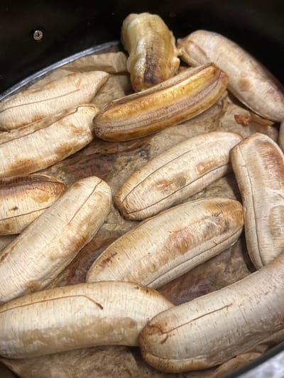 This close-up, overhead image displays numerous whole, unpeeled bananas or plantains arranged on a sheet of crumpled light brown parchment paper. The paper lines a dark, possibly metallic, cooking tray or container, the rim of which is visible along the left and bottom edges, along with a small silver screw in the lower-left corner.
The bananas are a pale, yellowish-beige color with varying degrees of golden-brown caramelization and browning on their skins, indicating they have been cooked. Some bananas exhibit more pronounced darker streaks and patches, particularly on their exposed surfaces. Most maintain their elongated, slightly curved shape. One banana on the far left appears partially peeled or burst open, revealing its softer, cooked, light yellow flesh.
The overall scene suggests the bananas are either in the process of being roasted or baked, or have just finished cooking. Their arrangement is somewhat informal, with some overlapping. There are no visible people, activities beyond the state of the food, or discernible text. The setting implies a kitchen or food preparation environment, with the specific location context of Cần Thơ, Vietnam, hinting at a local culinary preparation such as chuối nướng (grilled bananas).