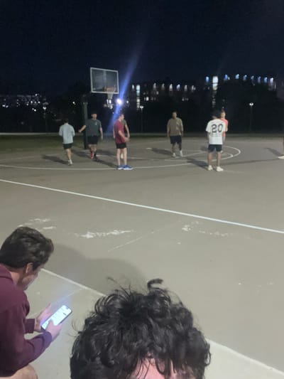 The image depicts an outdoor basketball court scene at night in College Station, United States of America.
In the immediate foreground, the top of a person's head with dark, curly hair is visible on the right. To the left, a second individual, wearing a maroon hooded sweatshirt, is holding and observing a brightly illuminated smartphone.
Seven individuals are present on the grey-surfaced basketball court, which features white boundary lines showing visible wear. From left to right on the court: one individual in a light grey shirt and dark shorts is walking to the left; one individual in a dark green shirt, dark shorts, and red shoes is standing centrally; one individual in a maroon shirt, black shorts, and blue shoes is holding a basketball; one individual in a white shirt with the number "20" on the back, dark shorts, and white shoes is standing; one individual in a light tan shirt, dark shorts, and white shoes is standing; and two additional individuals are partially visible on the far right edge of the frame, one wearing a light pink shirt.
A basketball hoop with a transparent backboard and rim is positioned on the left side of the court, illuminated by a strong floodlight located directly behind it, which produces a lens flare. Beyond the court, a dark ground expanse transitions to a distant background of multiple multi-story buildings with illuminated windows, consistent with institutional or residential architecture found on a university campus. Several illuminated streetlights are also visible, and a faint urban skyline is discernible on the far left horizon.