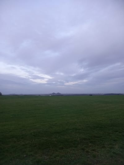 A vast green field covers the right half of the image. Overcast skies fill the left, with subtle clouds. The horizon line is nearly centered. A small, distant hill is visible near the horizon. A few scattered trees are also visible along the horizon, but they are dwarfed by the vast field and sky.