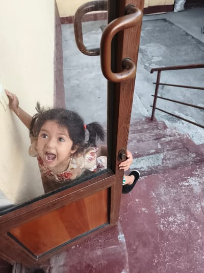 A young girl with dark hair in pigtails, wearing a light-colored dress with a floral pattern, is looking up with her mouth open as if calling out. She is seen through a glass door, her left hand is pressed against the wall, and her right hand is on the wooden door frame. Her body is angled towards the camera, suggesting she is on the other side of the door. The background shows a staircase with a reddish-brown, cracked surface, a metal railing, and a concrete floor. The lighting is dim. The overall scene appears to be an entrance to a building. Without further information, the specific location in Buz, Uzbekistan, cannot be determined from the image alone.
