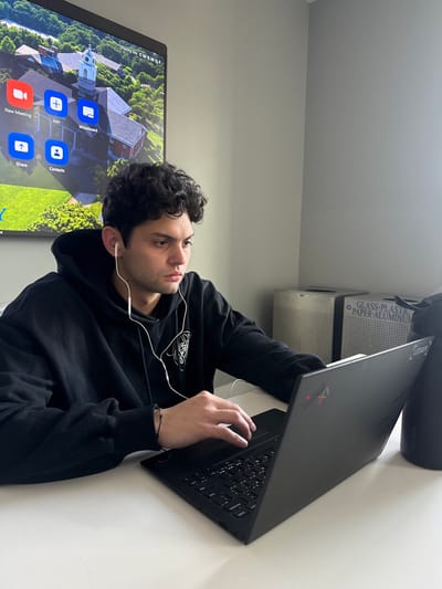 The image captures a young adult male seated at a white desk, actively using a black Lenovo ThinkPad laptop. He wears a black hooded sweatshirt with a white graphic design and white wired earbuds. A dark bracelet adorns his left wrist. His gaze is directed toward the laptop screen, with his hands positioned over the keyboard and trackpad. To his right, a black Yeti water bottle with a silver lid rests on the desk.
Behind the individual, a large wall-mounted flat-panel display shows the Zoom interface, featuring icons for "New Meeting," "Join," "Whiteboard," "Share," and "Contacts." The background image on this screen is an aerial view of Brandeis University in Waltham, Massachusetts, United States, highlighting a brick building with a prominent clock tower amidst green foliage. To the right, two grey, perforated recycling/waste bins are partially visible, one explicitly labeled for "GLASS-PLASTIC PAPER-ALUMINUM." The room's walls are painted a plain light grey.