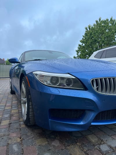 A blue BMW Z4 (E89 generation, likely post-facelift with M-Sport styling) is centrally positioned, viewed from a low-angle front-right perspective, in Куликово, Russia. The entire visible exterior of the vehicle, including the hood, windshield, and front fender, is covered in numerous small, spherical water droplets, consistent with recent rainfall or moisture exposure. The front right headlight, featuring dual projector lenses and illuminated LED corona rings, is prominent. The right half of the BMW kidney grille, characterized by vertical silver slats, is partially visible alongside the lower section of the M-Sport front bumper, which incorporates black mesh grilles and a rectangular cover for a tow hook on the right. The front right silver multi-spoke alloy wheel and its Michelin-branded tire are in focus, resting on the ground. The vehicle is parked on a wet, multi-colored interlocking paver driveway, which shows scattered organic debris. The background features an overcast sky with diffuse cloud cover. A large green deciduous tree occupies the upper right background. A portion of a white SUV is visible behind and to the right of the BMW. A dark metal fence and a section of a building with a dark roof are discernible in the far left background. No individuals are present in the image, and the vehicle is stationary.