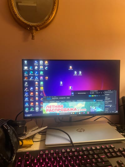 The image shows a desk setup with a computer monitor, keyboard, and laptop. The monitor displays a desktop with various icons and a wallpaper of a purple and pink gradient. A Steam browser window is open on the monitor, showing a sale advertisement. A black gaming keyboard with pink backlighting is in the foreground. A silver HP laptop is placed on the desk below the monitor. There is a black leather headphone case on the left side of the desk. The background features a wall with a large ornate gold mirror hanging above the desk. The overall environment appears to be an indoor personal workspace. There is no indication of the location being Sainte-Marie-la-Mer, France.