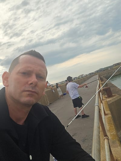 The image shows a man taking a selfie on a pier. In the background, a man is fishing, and two other people are visible. The location appears to be a coastal area with buildings in the distance. The sky is overcast with clouds.
