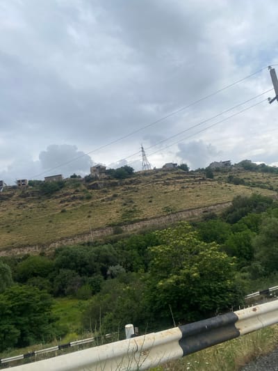 This image depicts a hillside with several houses scattered across it, a power line tower, and a guardrail. The hillside is covered in dry grass and some trees. The sky is cloudy, suggesting it might be a dreary day. The houses appear to be in various states of construction or disrepair. The overall scene is one of rural habitation in a hilly landscape. The guardrail is a prominent object in the foreground on the right side of the image, featuring a distinctive black and white striped pattern. The power line tower is visible in the lower left portion of the frame, with wires stretching across the sky. The image is oriented vertically, with the top of the frame showing the sky and the bottom showing the ground. The location is identified as Ptghni, Armenia.