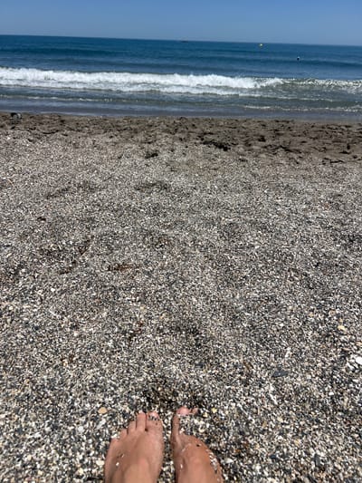 The image captures a perspective of a person's feet resting on a pebble beach, facing the Mediterranean Sea. The sand is a mixture of dark and light pebbles, with some damp patches closer to the water. Gentle waves are breaking on the shore, with a clear blue sky overhead. In the distance, a few buoys are visible in the sea, suggesting a marked area. The scene is peaceful and evokes a sense of relaxation on a sunny day. The location is implied to be a beach in San Roque, Spain.