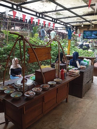 The image depicts an outdoor food stall area located in Kajar, Indonesia. In the foreground, a long, dark-wood food preparation and serving counter is positioned on a concrete floor under a white, translucent corrugated roof supported by dark metal beams. A string of red and white pennant flags hangs from the roof structure.

On the left side of the counter, a woman with a light-colored hijab and dark long-sleeved top is seated, facing towards the viewer but looking slightly to the left. In front of her on the counter, a large lidded pot, a colander, and several bowls containing prepared dishes (including some with light blue contents) are visible. Further along the counter to the right, numerous bowls of various prepared dishes are arranged, along with two red condiment bottles and a single white bottle. A large, dark wooden box or cabinet sits on the counter behind these items. To the right of this primary counter, another dark wooden counter or cabinet is present, topped with what appears to be packaged items. Behind this, a person wearing a dark blue hijab is partially visible.

In the midground, behind the food counter, there is a sloped, densely vegetated area with green plants and some larger trees. Two individuals are walking uphill in this area; one wears a light-colored shirt and pants, and the other wears a dark top and pants. To the far right of the scene, a flat-screen television is mounted, displaying what appears to be security camera footage. Below the TV, a black scooter is parked on the vegetated slope. Further back, several rustic-looking structures, possibly thatched huts or small buildings, are partially visible amidst the greenery.