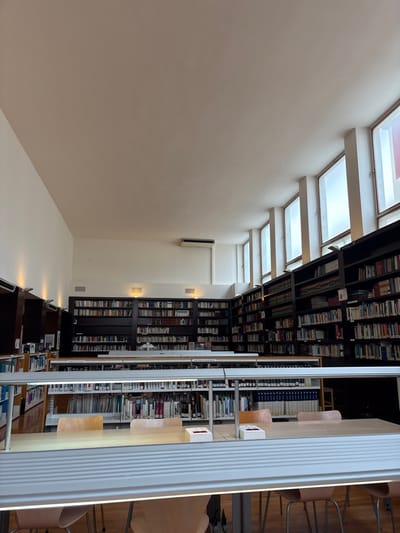 This image depicts the main reading room of the Biblioteca Pública Municipal d'Alcoi (Public Municipal Library of Alcoy) in Alcoi, Spain. The room features a high, flat, light-colored ceiling and smooth, light-colored walls. Along the upper section of the right wall, a series of seven tall, narrow, rectangular windows with frosted or translucent panes allow natural light to enter. A white split-system air conditioning unit is mounted high on the back wall.
Extensive dark-colored bookshelves filled with numerous books line the back and right perimeter walls, extending upwards to a significant height. In the foreground and midground, multiple long, light-colored wooden study tables are arranged parallel to the bookshelves. Each table is equipped with an integrated horizontal fluorescent lighting fixture suspended directly above its surface. Several light-colored wooden chairs are positioned at these tables; two are clearly visible in the immediate foreground, partially obscured by the lighting fixture, and others are visible further back. Some tables feature small, white, rectangular power modules or outlets. The flooring appears to be made of light-colored wood. No individuals are present within the scene.