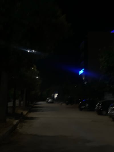 The image shows a dark, nighttime scene, likely in an urban setting. A row of cars is parked along the right side, near what appears to be a sidewalk. In the distance, there are faint lights. The image suggests an environment where lighting is dim, typical of a residential area in the evening. Specific location within Dar Oum Soltane, Morocco, is unidentifiable.