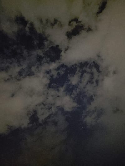 The image displays a dark, cloudy night sky over Mladá Boleslav, Czechia. Dense, textured cumulus clouds, varying in shade from light grey to dark grey, dominate the frame, obscuring most celestial features. Scattered patches of dark blue-black sky are visible through breaks in the cloud cover, particularly towards the center and lower right. Faint, pinprick-like white points, indicative of stars, are discernible within these clearer sky sections. The overall scene is dimly lit, suggesting ambient light reflecting off the clouds or minimal natural illumination. The image exhibits significant visual noise, consistent with a low-light photographic capture. No human subjects, man-made structures, or explicit actions are present within the frame; the subject is exclusively the atmospheric conditions.