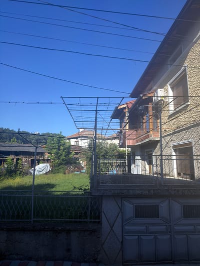 In Hadzhidimovo, Bulgaria, a residential scene unfolds under a clear blue sky. The foreground is dominated by a grey metal fence with an ornate scrolling pattern, atop a concrete base. Beyond the fence, a driveway gate with a segmented design is visible.
The middle ground features a small yard with lush green grass and scattered yellow wildflowers. A white tarp is draped over an object in the yard, and a rustic wooden structure with a dark, weathered roof stands to the left. A metal trellis system, seemingly for vines or climbing plants, extends across part of the yard.
To the right, a two-story building with a textured, yellow-speckled facade rises. It has a balcony with wooden railings and several windows, some covered with brown shutters. An outdoor air conditioning unit is mounted on the side of the house. Another building with a red-tiled roof is visible behind it.
The background consists of a dense line of green trees, with a faint white structure perched on a distant hill. Power lines crisscross the bright sky, a common sight in many residential areas. The overall impression is one of a quiet, rural or semi-rural Bulgarian setting on a sunny day.