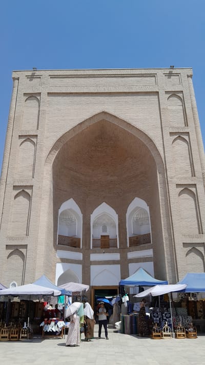 In Denov, Uzbekistan, a large, imposing brick building with an arched entrance dominates the frame. The architecture features multiple recessed arches and geometric patterns in the brickwork. In front of the building, a bustling marketplace is set up with stalls covered by colorful awnings. Several people are present, some browsing goods displayed on tables and racks, while others are walking or standing. The marketplace appears to be selling textiles, clothing, and decorative items. The sky is a clear, bright blue.