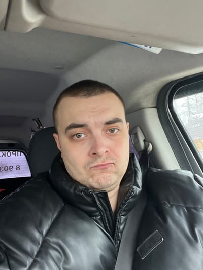 The image displays an adult male seated in the front passenger or driver's seat of a vehicle, located in the city of Rogachovo, Russia. The individual, appearing to be in his 30s-40s, has short dark hair, light stubble on his face, and brown eyes. He is wearing a black puffer jacket and a seatbelt is visible across his chest. His facial expression indicates a slight pout or concern, with subtly furrowed eyebrows. The vehicle's interior features a grey headliner and grey plastic trim around the window frame. Through the rear window, partially obscured by reflections, Cyrillic text fragments "ШPOK" and "e 8" are visible on an external object. The view through the side window on the right shows a blurry outdoor environment with bare tree branches against an overcast sky, suggesting a non-summer season. The man is looking directly towards the camera, suggesting a selfie or direct photograph.