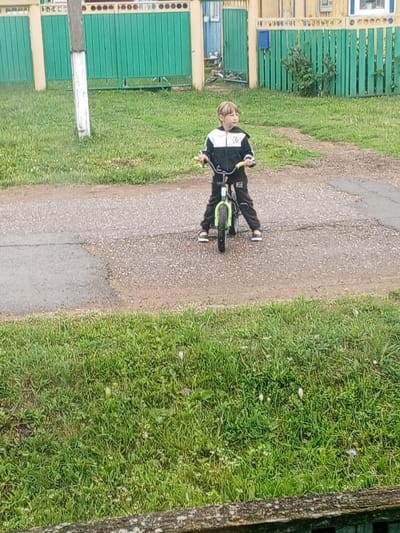 In Buraly, Russia, a young boy with blond hair is riding a green bicycle on a paved path. He is wearing a black and white tracksuit and white sneakers. The boy is looking to his right and appears to be focused on something off-camera. The background features a green wooden fence with a white pole in front of it, and some houses with yellow and blue accents. The ground is a mixture of asphalt and gravel, with a grassy area on the left and foreground. The weather appears to be overcast.