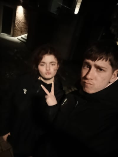 Two individuals, a male and a female, are captured in a selfie taken at night. The female is on the left, with brown, curly hair, making a peace sign. She is wearing a dark jacket with a visible Nike logo. The male is positioned to the right and has short, dark hair, with a somewhat stern expression. He is also wearing a dark jacket. The background includes a building lit by an artificial light source, revealing a brick wall and steps. The image suggests an urban environment, potentially Emmerich on the Rhine, Germany. The lighting conditions are dim, indicating it is likely nighttime. The subjects are close to the camera.