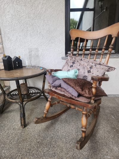 A vintage wooden rocking chair is placed on a granite tile floor, possibly on a porch or patio. The chair is adorned with several cushions, including one with a patterned beige cover, a light blue one, and two in muted purple and brown tones. To the left of the chair, a small, round wicker table with a glass top holds two miniature black house decorations and a clear glass vase. The background features a textured white wall and a window. The scene suggests a relaxed outdoor setting.