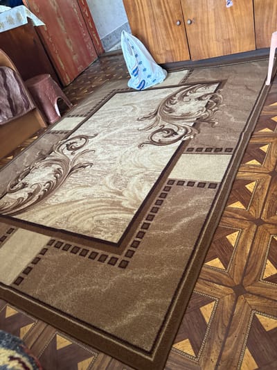 The image depicts an indoor scene within the city of Tihomir, Bulgaria. The floor features a large, rectangular rug predominantly in shades of brown and beige. The rug has a central beige panel adorned with dark brown ornate, swirling patterns, bordered by a darker brown section with a series of small squares, and an outermost solid dark brown edge. Adjacent to the rug, the exposed floor is covered with patterned linoleum or parquet flooring, exhibiting a repeating geometric design of dark and light wood-toned triangles and squares, with thin, woven-like borders around the lighter sections.
Along the left side of the frame, a light brown wooden bed frame or sofa base is partially visible, topped with a brown and beige striped fabric. A reddish-brown plastic stool, resembling a tree stump with an arched base, rests on the floor beside it. Further back, two reddish-brown wooden wardrobe doors with vertical grain are visible. On the right side, a larger wooden wardrobe with a lighter brown hue and two doors, each featuring a silver-colored knob, stands against a light-colored wall. A small, light pink plastic stool leg is partially visible on the far right.
A clear or white plastic shopping bag, displaying blue text and logos in Cyrillic script, is positioned on the rug near the upper center of the image. No people are present, and no specific actions or interactions are depicted.
