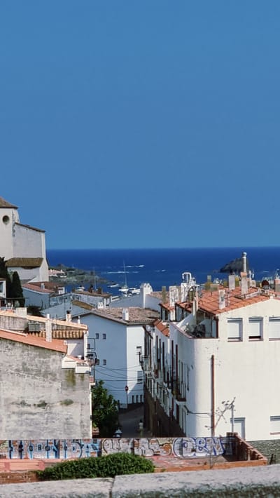 The image shows a view of the town of Cadaqués, Spain, overlooking the Mediterranean Sea. The foreground is dominated by the rooftops of white buildings with terracotta tiles, characteristic of Mediterranean architecture. Some buildings have balconies and numerous chimneys are visible. In the middle ground, more white buildings with red-tiled roofs line a narrow street, with a few trees and a single person walking. The background features the deep blue of the sea, dotted with several sailboats and a distant lighthouse on a rocky outcrop. The sky above is a clear, vibrant blue. The overall scene depicts a sunny day in a coastal European town.