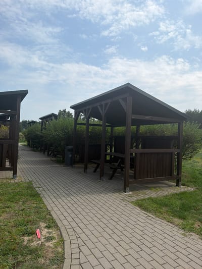 The image depicts an outdoor scene with multiple wooden gazebos lined up along a paved walkway in the city of Polesie, Poland. The gazebos are dark brown and appear to be of a similar design, each with a roof, supporting posts, and slatted wooden walls. Inside some of the gazebos, benches and tables are visible. A dark grey trash can is situated next to one of the gazebos. The walkway is paved with light-colored rectangular bricks and is bordered by green grass on one side. Behind the gazebos, dense green bushes and trees form a natural backdrop, with a hint of a forest in the distance. The sky above is partly cloudy with scattered white clouds against a light blue background. The overall scene suggests a recreational or picnic area, possibly within a park or campground. There are no people visible in the image, and no overt actions or interactions are taking place. The environment appears to be natural and maintained. A small red object, possibly litter, is visible on the grass near the walkway in the foreground.