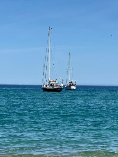 The image captures a tranquil scene at sea, likely in the vicinity of Le Lavandou, France. Two sailboats are anchored in the clear, turquoise waters under a vast, cloudless blue sky. The sailboat in the foreground is dark-hulled with tall masts, while the second sailboat, slightly further back, is lighter-colored. The sea exhibits gentle waves, and the horizon line is visible in the distance, separating the blue of the water from the sky. There are no people visible in the image, and the sailboats appear to be at rest, suggesting a peaceful day. The overall impression is one of serene maritime beauty.
