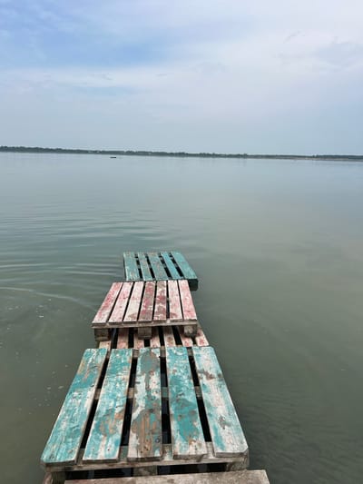 This is a photograph taken from an eye-level perspective looking out over a calm body of water. In the foreground, a makeshift pier constructed from weathered wooden pallets juts into the water. The pallets are stacked and have planks laid across them, some painted with chipped turquoise and red paint. The water is a muted gray-green, with gentle ripples indicating a light breeze. In the distance, a treeline marks the far shore, with a few buildings visible. The sky above is overcast with a soft, diffused light. This scene is located at the resort Lake Seliger in Russia.