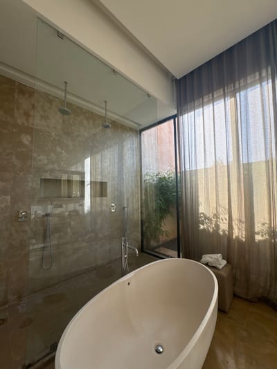 A bathroom located in a luxury villa in Ouled Jelal, Morocco (31.664, -7.87) features a large walk-in shower with a glass enclosure and two shower heads. The shower is tiled with brown marble-like tiles. A white, oval-shaped bathtub sits in front of the shower, filled with water. A window to the right of the shower reveals a small outdoor area with a courtyard and greenery. Sheer, brown curtains hang in front of the window. A white towel lies folded on a small, brown ottoman next to the bathtub. The floor is made of light brown tile. The overall decor is modern and minimalist, with a focus on sleek lines and natural materials. There are no people present in the room.