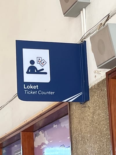 A blue sign with a white graphic is mounted on a textured wall. The graphic depicts a stylized person sitting and holding two playing cards, each with a downward-pointing arrow. Below the graphic, the sign reads "Loket" in larger white font and "Ticket Counter" in smaller white font. The sign is illuminated by an unseen light source, casting subtle shadows. To the right of the sign, a brown wooden door frame is visible. The wall behind the sign has electrical wires and two speaker-like fixtures attached to it. The overall environment appears to be an indoor public space, possibly a transportation hub or a public building in Desa Tambakreja, Indonesia.