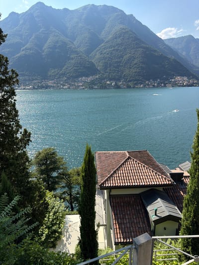 A bright, sunny, vertical, eye-level shot captures a picturesque landscape of Lake Como, Italy, viewed from Pognana Lario. The lake dominates the center of the frame, its surface shimmering with sunlight and reflecting the deep blue sky. In the distance, the verdant, tree-covered slopes of mountains rise majestically, dotted with a cluster of white buildings that form a small village. The lower portion of the image showcases the foreground, featuring the roof and exterior of a building with red-tiled roofing. Lush green foliage, including tall cypress trees, frames the scene, adding depth and a sense of natural beauty. The overall impression is one of tranquility and scenic splendor.