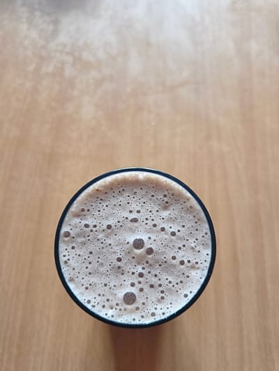 A top-down shot shows a foamy, light-brown beverage in a black-rimmed glass on a light-brown wooden surface. The drink is covered with numerous small bubbles, with a few larger bubbles dispersed throughout. The wooden surface appears to be a table or countertop. The location is unknown but is likely in Russia. Kstovo is a city that is on the Volga River. The city is known for its petrochemical and oil refining industries.