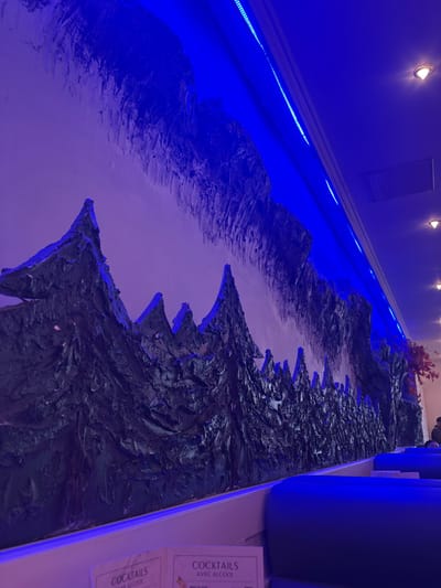 The image depicts the interior of a commercial establishment, likely a restaurant or lounge, located in Audun-le-Tiche, France. A prominent 3D relief mural covers a significant portion of the main wall, illustrating a stylized mountainous landscape with jagged peaks and textured evergreen trees in dark blue or deep green tones. Above the treeline, a textured representation of rock formations or a sky is rendered in similar relief. A continuous blue LED strip light runs along the ceiling perimeter above the mural, casting a strong blue illumination onto the wall art and portions of the white ceiling. The ceiling also features multiple recessed spotlights emitting warm, yellow-white light. In the foreground and extending into the midground, several blue upholstered booth seats are arranged parallel to the wall, separated by a white ledge. On this ledge, two white menu cards are partially visible, both displaying "COCKTAILS" and "AVEC ALCOOL." In the far right background, a decorative plant with reddish-brown foliage is present. A single individual, clad in a dark top, is partially visible in the extreme right background, obscured by the plant and seating. No specific actions or interactions are clearly discernible from this individual.