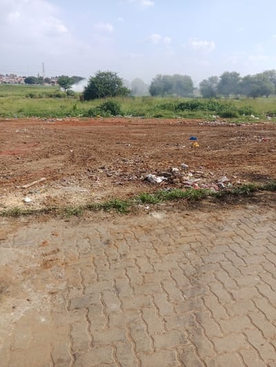 A wide shot shows a litter-strewn vacant lot adjacent to a paved, interlocking brick area. Garbage is concentrated in the middle of the lot, with sparse debris scattered elsewhere. Beyond the lot, a grassy area with trees stretches into the distance where smoke can be seen. Buildings and a utility tower are visible on the horizon under a clear, blue sky.