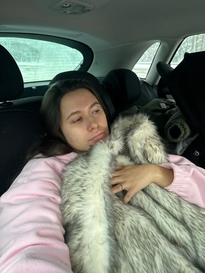 A female subject is depicted reclining in the rear passenger seat of a vehicle in Selyatino, Russia. She has long, dark brown hair and is wearing a light pink long-sleeved garment. She is holding or wrapped in a grey and white faux fur blanket, with her right hand visible on the blanket, displaying a simple gold-colored ring on her left ring finger. Her gaze is directed upwards and to the right, exhibiting a neutral to slightly pensive expression. The car interior features dark headrests on the front seats and a light-colored ceiling with a central light fixture. Through the rear windows, a blurry outdoor scene consistent with a winter landscape, possibly snow-covered, is visible. A dark green jacket and a portion of a dark car seat or similar object are present on the seat to the subject's right.