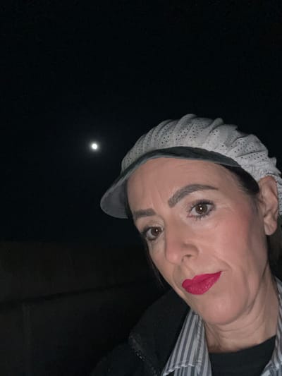 A forensic examination of the image reveals a nighttime outdoor scene in Roodt-sur-Syre, Luxembourg. The primary subject is an adult female, positioned in the lower right quadrant of the frame, angled towards the camera in what appears to be a self-portrait. She is wearing a light-colored, possibly white or light grey, perforated or textured cap with a brim, dark hair pulled back beneath it. Her facial features include defined eyebrows, brown eyes, and bright red lipstick. She is attired in a light-colored, likely striped, collared shirt under a dark jacket or coat. The background consists of a dark, unlit sky, with a visible, illuminated moon positioned in the upper left quadrant of the image. The immediate environment behind the individual is indistinct and appears devoid of artificial light sources or discernible structures, except for a faint, dark horizontal line along the bottom edge of the distant background. No other individuals, objects, or specific actions beyond posing are observable.