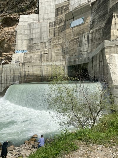 The image captures the Chirkey Dam, a significant infrastructure in Untsukul', Russia, characterized by its immense concrete structure and the powerful flow of water. The dam's face is marked with various inscriptions and graffiti, some in Cyrillic script. A section of the dam features an opening, likely a tunnel or spillway, through which water is visibly flowing.
In the foreground, a portion of the riverbank is visible, with scattered rocks and lush green grass. Two individuals are present near the water's edge. One person, dressed in dark clothing, stands with their back to the viewer, looking towards the dam. Another individual, wearing a blue and white striped shirt, is seated on the rocks, facing the water. The surrounding environment suggests a natural setting, with a rocky cliff face adjacent to the dam. The sunlight illuminates the scene, casting shadows and highlighting the textures of the concrete and the water.